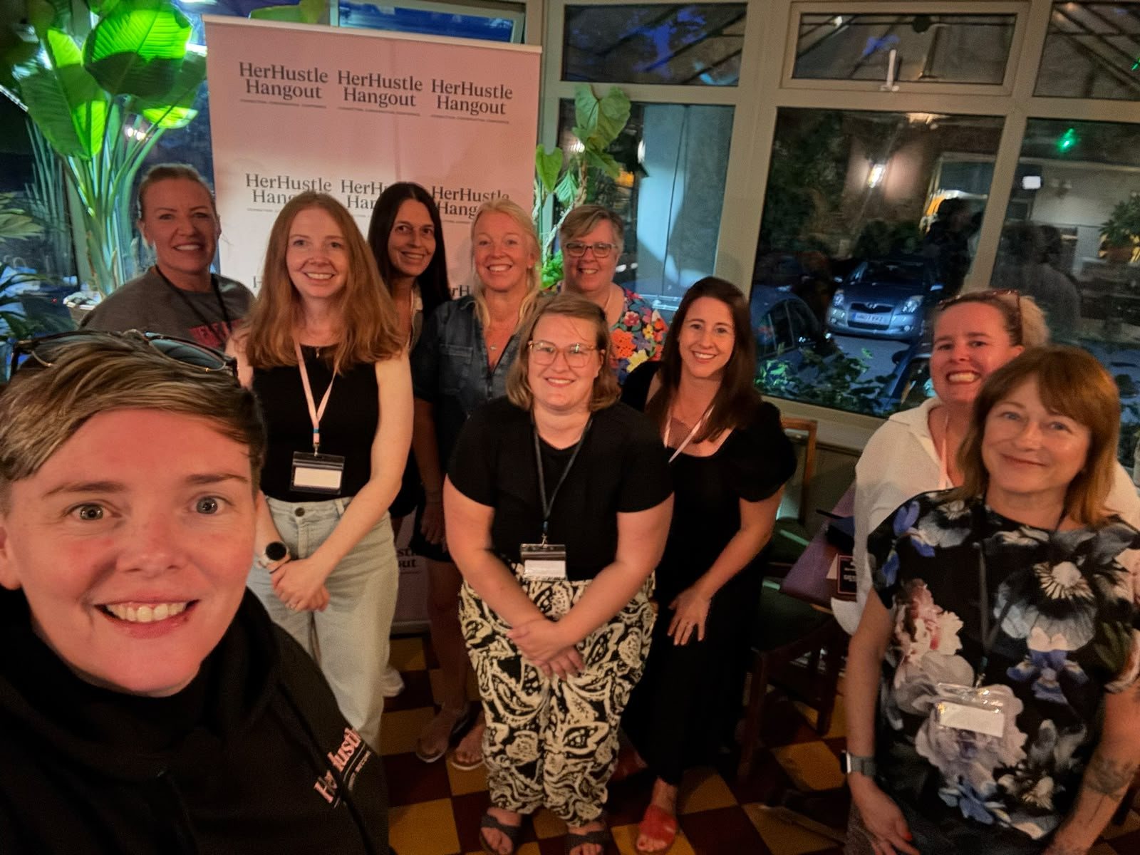 Ten women smiling in a group selfie in front of a HerHustle Hangout banner.