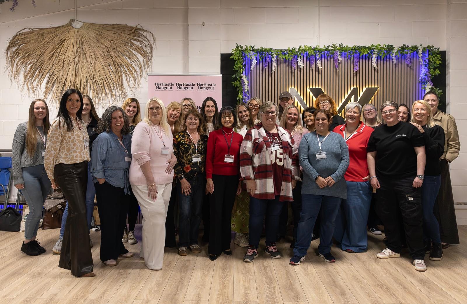 A large group of women smiling together in front of a HerHustle Hangout banner.