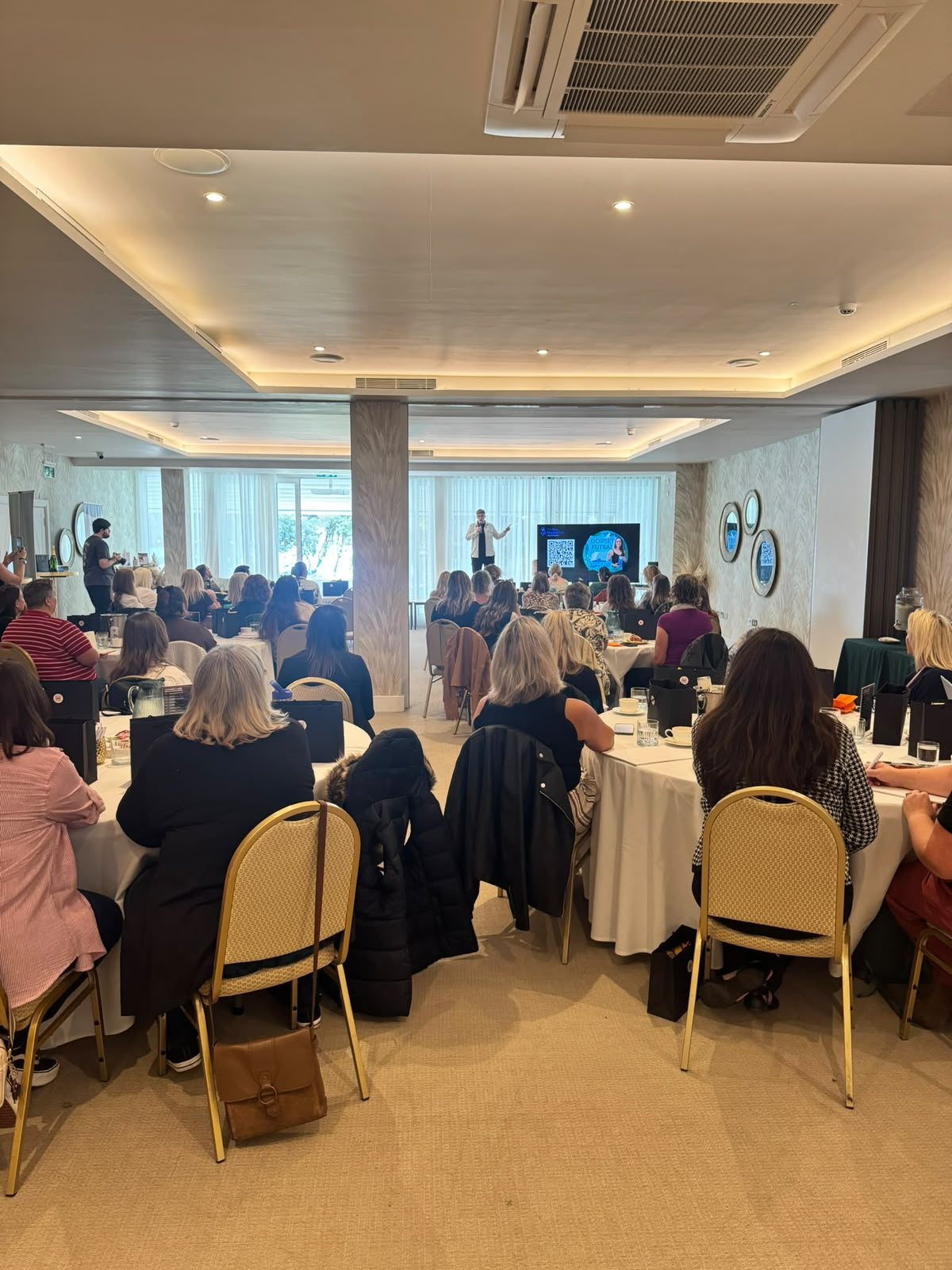 A packed room of women at round tables, watching the speaker at the front of a HerHustle event.