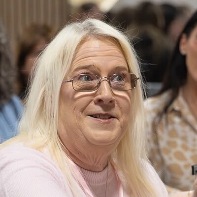 Jo Shelton, with long blonde hair and glasses, speaking from her seat at a HerHustle event.