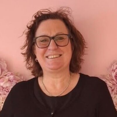 Julie Miller, smiling in glasses and a black cardigan in front of a wall of pink flowers.